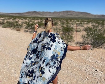 Può includere: Una donna indossa un kimono a stampa floreale bianco e blu con un bordo blu scuro. Il kimono fluttua nel vento mentre cammina attraverso un paesaggio desertico.