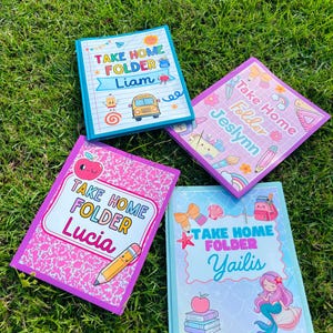 May include: Four rectangular folders labelled Take Home Folder with personalised names Liam, Jeslynn, Lucia, and Yailis. The folders feature colourful cartoon graphics including a school bus, school supplies, a mermaid, and patterned backgrounds.
