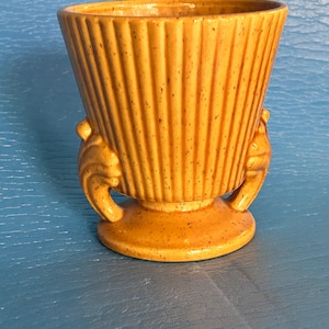 Puede incluir: Una maceta de cerámica vintage en un cálido tono naranja moteado. La maceta presenta detalles acanalados verticales y dos asas decorativas. Tiene una base redonda y una parte superior abierta y ensanchada, adecuada para plantas pequeñas o arreglos florales.