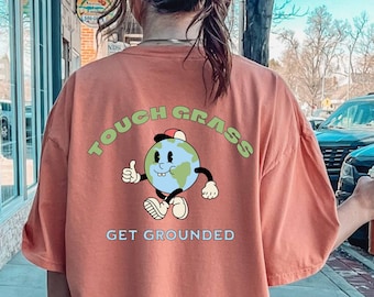Puede incluir: Una camiseta de color coral con una ilustración de dibujos animados de una tierra sonriente con brazos y piernas. La tierra lleva una gorra de béisbol roja y hace un pulgar hacia arriba. El texto "Touch Grass Get Grounded" está impreso en un círculo alrededor de la tierra.