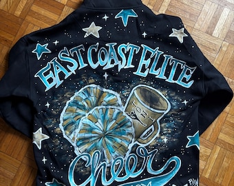 May include: Black zip-up jacket with hand-painted design. Features the text "East Coast Elite" and "Cheer Mom" in teal and white. Includes illustrations of pom-poms and a coffee mug, with star accents.