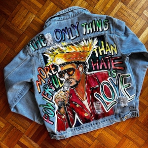 Puede incluir: Chaqueta vaquera azul claro con un diseño pintado colorido. La obra de arte presenta a una persona cantando en un micrófono, con las palabras "THE ONLY THING MORE POWERFUL THAN HATE IS LOVE" en letras vibrantes. La chaqueta tiene detalles desgastados.