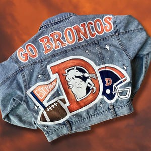Puede incluir: Chaqueta vaquera azul claro con texto "GO BRONCOS" y logotipos del equipo pintados a mano. Presenta una gran "D" naranja con un caballo blanco, un balón de fútbol y un casco. La chaqueta incluye el texto "EST. 1960" en una bandera.