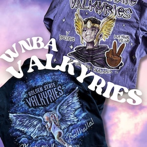Könnte beinhalten: Zwei Hemden mit handgemalten Designs. Eines ist lila mit "Golden State Valkyries" und einer geflügelten Figur. Das andere ist schwarz mit "Golden State Valkyries" und einer ähnlichen Illustration. Das Thema ist das WNBA-Team.