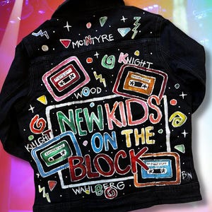 May include: Black denim jacket with colorful hand-painted graphics. The jacket features the words "New Kids on the Block" and various cassette tape designs. Names like "McIntyre", "Knight", "Wood", and "Wahlberg" are also visible.
