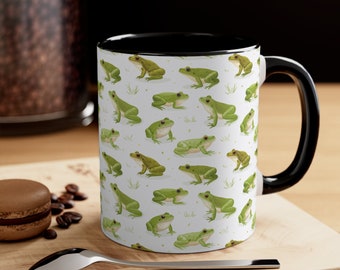 May include: A white ceramic mug with a black rim and handle. The mug features a repeating pattern of green frogs on a white background.