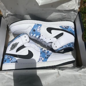 May include: Pair of white and light blue high-top trainers with black detailing. The trainers feature a basketball logo and the words "NO CA" and "UNC" on the sides. They are presented in a box with white tissue paper.
