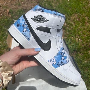 May include: White and blue Air Jordan 1 Mid trainer with a black Nike swoosh. The shoe features a blue pattern with cartoon figures and text. The Air Jordan logo is visible on the side.