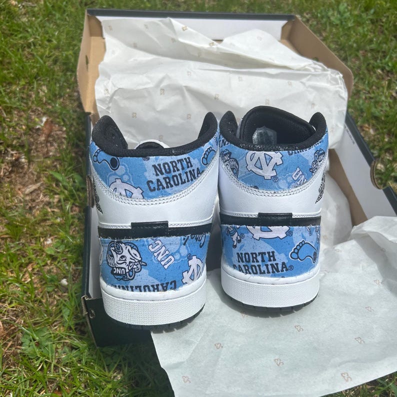 May include: Pair of white and light blue high-top trainers with "NORTH CAROLINA" printed on the back. The trainers feature a light blue pattern with the UNC logo and other graphics. The shoes are in an open shoe box.