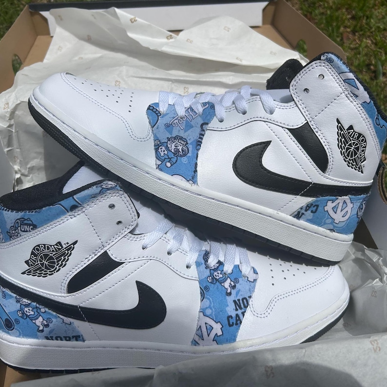 May include: White and black Air Jordan 1 Mid trainers with light blue detailing. The trainers feature the Air Jordan logo, a black Nike swoosh, and light blue patterned panels with text and cartoon figures. The shoes are in a cardboard box.