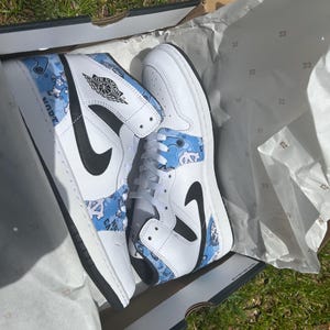 May include: White and blue high-top trainers with black detailing, showcasing a distinctive pattern and the Air Jordan logo. The trainers are presented in a box with tissue paper.