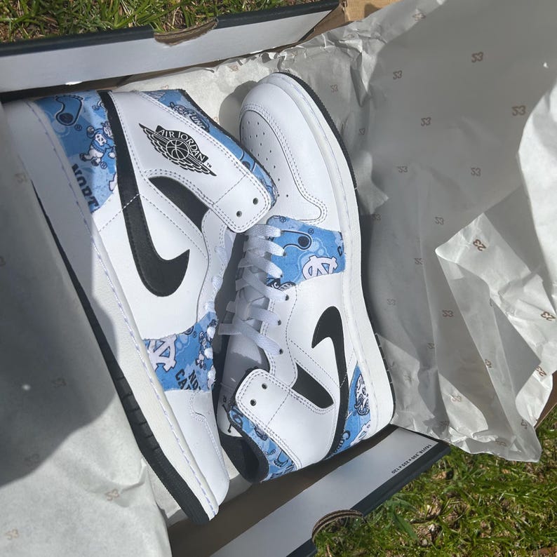 May include: White and blue high-top trainers with black detailing, showcasing a distinctive pattern and the Air Jordan logo. The trainers are presented in a box with tissue paper.