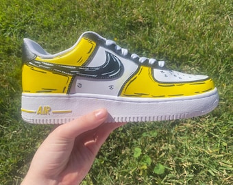 May include: Custom-painted white and yellow sneakers with a black Nike swoosh and black cartoon-style outlines. The sneakers have the word "AIR" printed on the side.