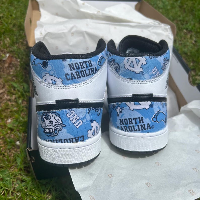 May include: Pair of white and light blue high-top trainers. The trainers feature a light blue pattern with the words "North Carolina" and "UNC" printed on the sides. The shoes are in a box.