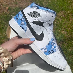 May include: White and black Air Jordan 1 Mid trainer with a black Nike swoosh. The shoe features light blue patterned panels with cartoon figures and the words "North Carolina" and "UNC". The Air Jordan logo is visible on the side.