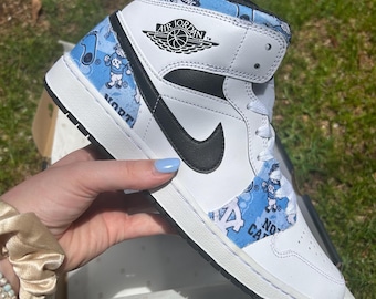 Puede incluir: Zapatilla Air Jordan 1 Mid blanca y negra con un logotipo de Nike negro. La zapatilla presenta paneles estampados en azul claro con figuras de dibujos animados y las palabras "North Carolina" y "UNC". El logotipo de Air Jordan es visible en el lateral.
