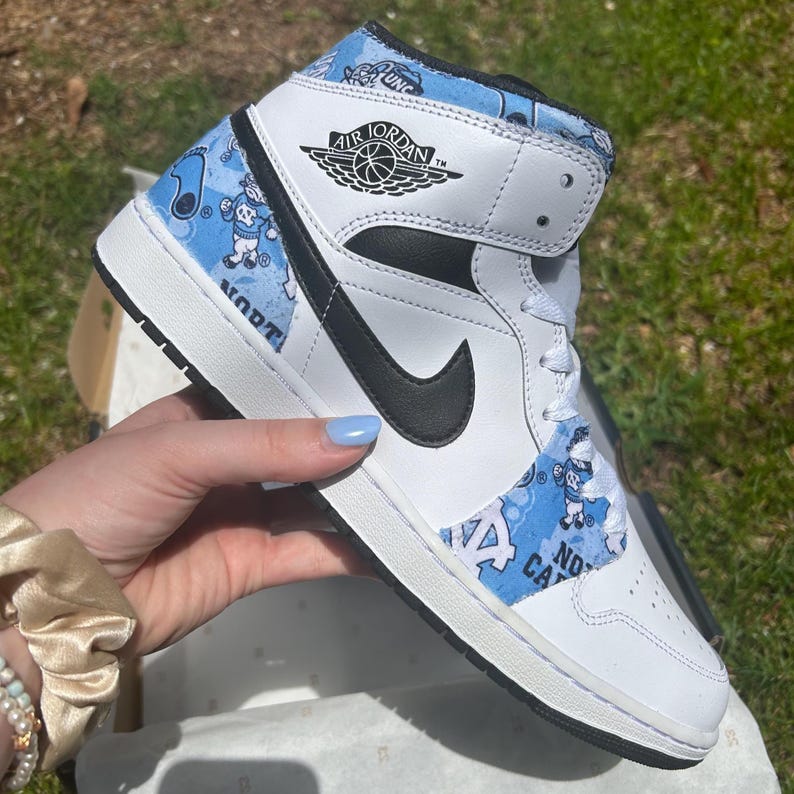 May include: White and black Air Jordan 1 Mid trainer with a black Nike swoosh. The shoe features light blue patterned panels with cartoon figures and the words "North Carolina" and "UNC". The Air Jordan logo is visible on the side.