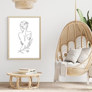 May include: A black and white line drawing of a woman's back with flowers, framed in a light wood frame, hanging on a white wall. A wicker hanging chair with a white cushion and throw blanket is in the foreground.