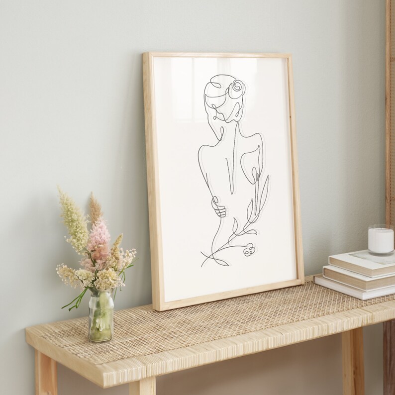 May include: A framed black and white line drawing of a woman with a flower in her hair. The drawing is on a white background and is framed in a natural wood frame. The frame is sitting on a light brown wooden table with a woven rattan top.