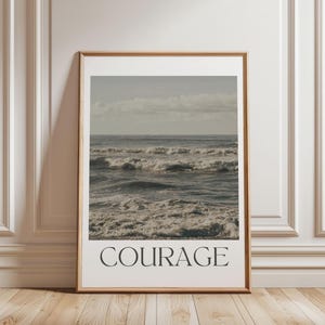 Puede incluir: Una impresión enmarcada con una fotografía de olas del océano rompiendo en una playa. La impresión tiene la palabra "COURAGE" impresa en negro en la parte inferior.