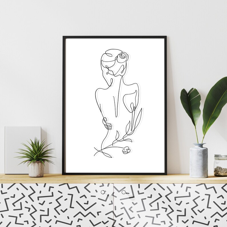 May include: Black and white line art print of a woman with a flower in her hair. The print is framed in a black frame and is hanging on a white wall.