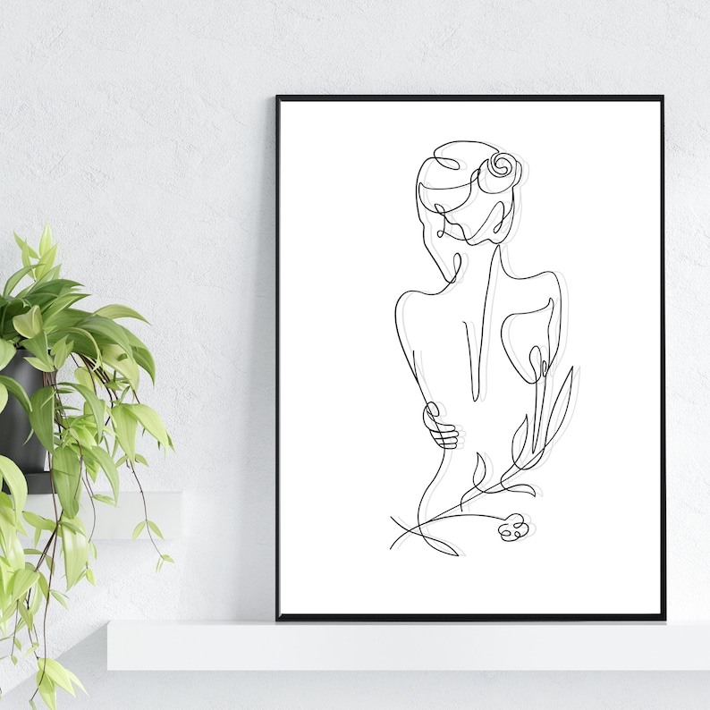 May include: Line art drawing of a person with a flower in their hair, standing with their back to the viewer. The drawing is in black and white and is framed in a black frame.