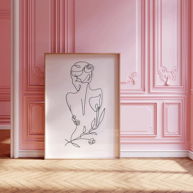 May include: A line drawing of a person with a flower in their hair, in a minimalist style. The drawing is in black and white and is framed in a gold frame. The frame is hanging on a pink wall.