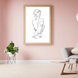 May include: A black and white line drawing of a person with a flower in their hair. The drawing is framed in a light wood frame and is hanging on a pink wall.
