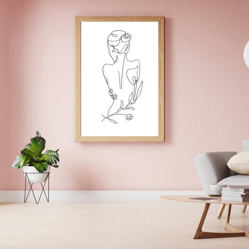 May include: A black and white line drawing of a person with a flower in their hair. The drawing is framed in a light wood frame and is hanging on a pink wall.
