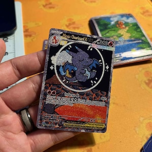 May include: A Mega Charizard X Pokemon trading card. The card features a purple Charizard illustration within a circular frame, with the text "Mega Charizard X" and "Inferno X". The card has a black background with red and orange accents.
