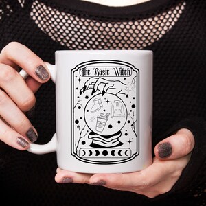 Puede incluir: Taza de cerámica blanca con una ilustración en blanco y negro de una mano de bruja sosteniendo una bola de cristal. El texto "The Basic Witch" está impreso en la taza.