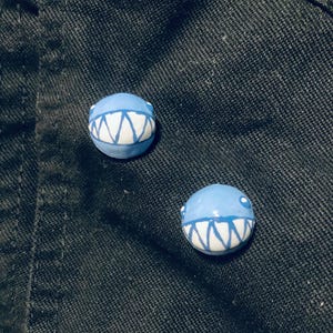 Peut inclure: Deux perles rondes en céramique bleu clair avec des motifs de dents blanches et dentelées. Chaque perle a un petit point blanc sur le côté, ressemblant à un œil. Les perles sont posées sur un fond sombre et texturé.