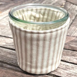 May include: A clear glass jar encased in a fabric sleeve featuring a vertical striped pattern in beige and off-white. The sleeve is stitched at the top rim and fits snugly around the exterior of the cylindrical glass container, providing a textured cover.
