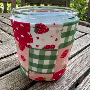May include: A glass jar wrapped in a fabric cozy featuring a patchwork design of red strawberries on white and pink backgrounds, interspersed with green and white gingham squares. The fabric cover has a stitched border along the top rim.