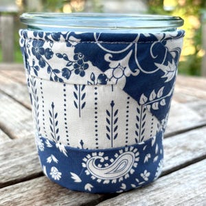 May include: A glass jar with a decorative fabric sleeve. The sleeve is made of blue and white patterned fabric, featuring floral, paisley, and dotted designs. The jar appears to be sitting on a wooden surface.
