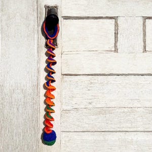 May include: A colorful crocheted toy with a spiral design and a ball at the end. The toy is hanging from a door handle.