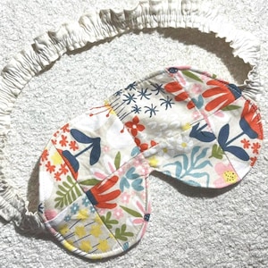 May include: A sleep mask featuring a colorful floral pattern with red, blue, pink, and yellow botanical motifs on a cream background. The mask includes a gathered, elasticized cream-colored fabric strap designed to secure the piece in place.