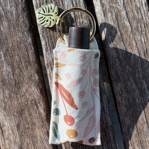 May include: A white fabric lip balm holder with a floral pattern in shades of pink, orange, and green. The holder is attached to a gold keyring with a gold leaf charm. A dark brown lip balm is inside the holder.