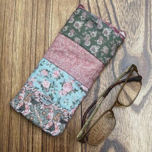 May include: A patchwork glasses case with floral patterns in shades of green, pink, and blue. Next to the case are gold-rimmed eyeglasses with rectangular lenses. The case is made of fabric and appears to be quilted.