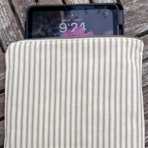 May include: A rectangular fabric sleeve featuring a cream and tan vertical striped pattern with a metal zipper closure at the top. A black tablet with a screen displaying the time 9:24 and the date Saturday, October 11 is partially inserted inside.
