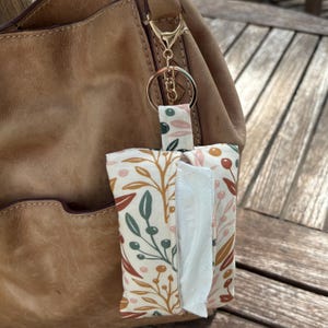May include: A floral print tissue holder with a gold-colored keyring attached to a brown leather bag. The fabric features a cream background with green, orange, and pink botanical designs. The holder is rectangular with a slot for tissue dispensing.
