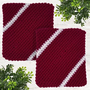May include: Two square, knitted potholders. The potholders are a deep burgundy color with a diagonal white stripe. The potholders are made of a thick, textured yarn. The potholders are displayed on a white surface with green foliage in the background.