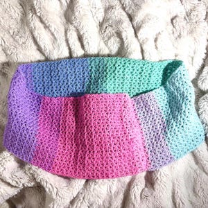 May include: A handmade crochet cowl in shades of purple, pink, blue, and green. The cowl is made with a open stitch pattern and is laying on a soft, textured surface.