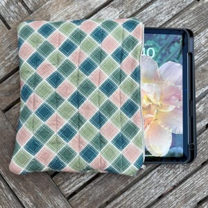 May include: A quilted tablet sleeve with a plaid pattern in shades of green, teal, and pink. The sleeve is open, revealing a tablet with a floral wallpaper. The sleeve is on a wooden surface.