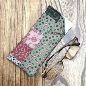 May include: A patchwork eyeglasses case with a floral pattern in shades of green, pink, and burgundy, next to a pair of gold-rimmed eyeglasses. The case is rectangular and appears to be handmade.