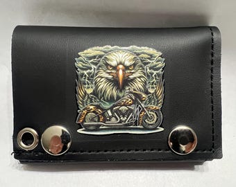 May include: Black leather wallet featuring a detailed graphic of an eagle with a motorcycle. The design includes a motorcycle with flames and an eagle with a stern expression. The wallet has silver snap closures.