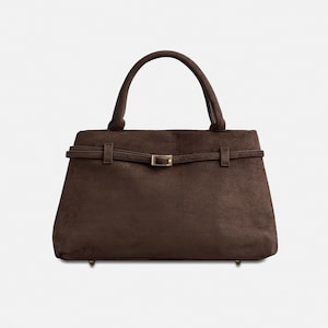 Puede incluir: Un bolso de mano de ante marr&oacute;n con asa superior y forma rectangular. El bolso presenta un detalle de cintur&oacute;n con hebilla dorada y peque&ntilde;os pies dorados. El bolso mide aproximadamente 30 cm de ancho.