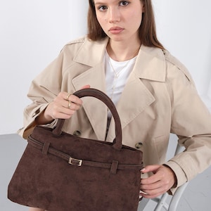 Puede incluir: Un bolso de mano de ante marr&oacute;n con forma rectangular y asa superior. El bolso presenta un detalle de cintur&oacute;n con una hebilla dorada. El bolso lo sostiene una persona que lleva una gabardina beige.