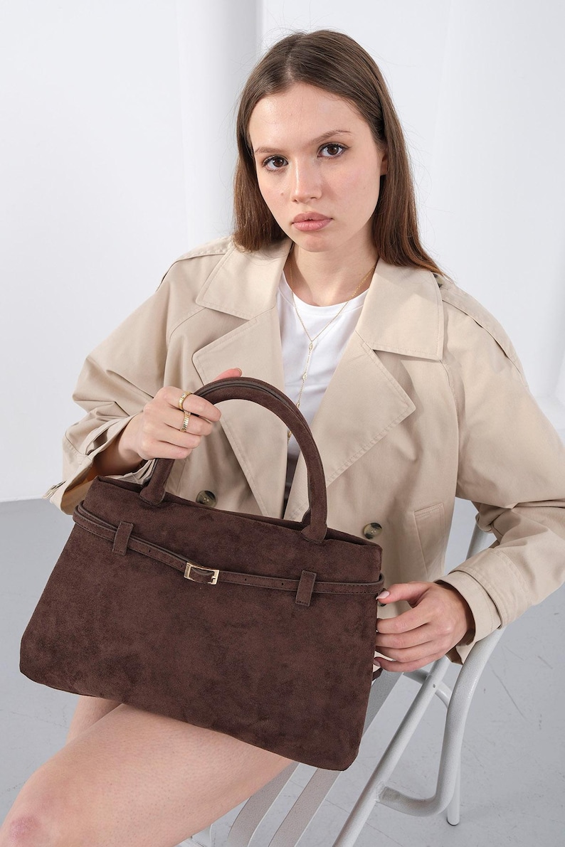 Puede incluir: Un bolso de mano de ante marr&oacute;n con forma rectangular y asa superior. El bolso presenta un detalle de cintur&oacute;n con una hebilla dorada. El bolso lo sostiene una persona que lleva una gabardina beige.