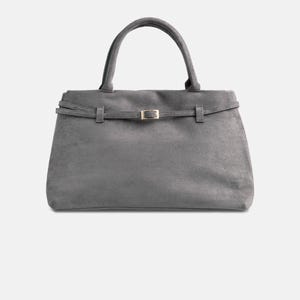Puede incluir: Un bolso gris con asa superior y detalle de cintur&oacute;n en la parte delantera. El bolso tiene forma rectangular con cierre de solapa y hebilla dorada. El material parece ser ante o un tejido suave similar.